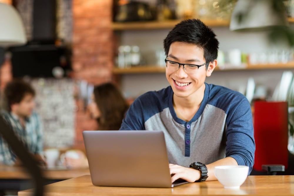 Happy cheerful young asian male in glasses smiling and using laptop in cafe - mudahnya komunikasi dengan pelanggan lewat aplikasi multiplechat